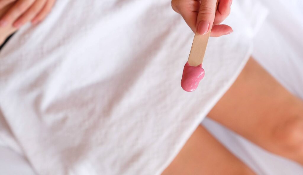 kvinde med hvidt håndklæde på lårene og close up af lyserød hot wax på spatel
