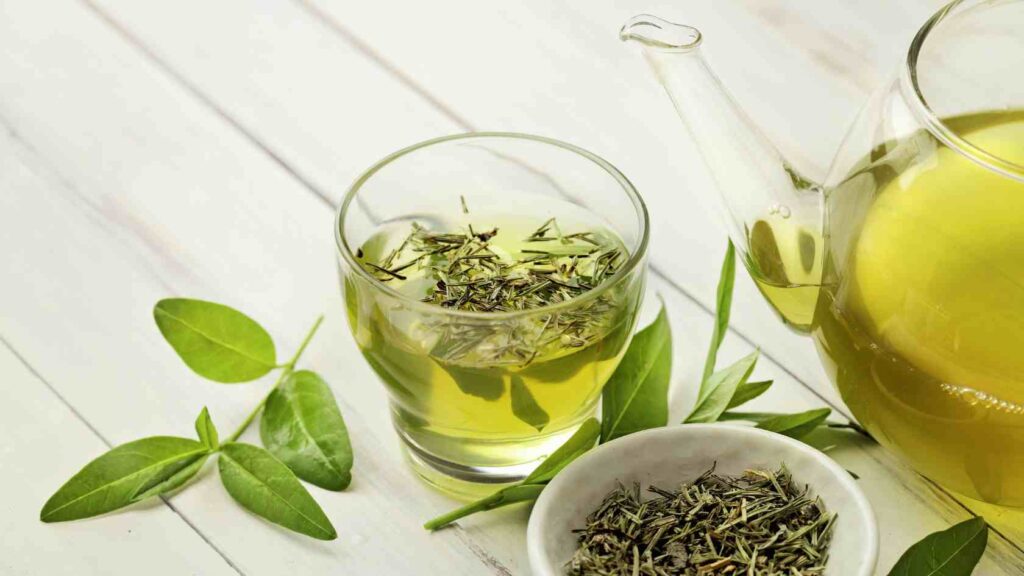 Groen te i glaskop med få blade rundt om koppen, samt en kande med groen te i